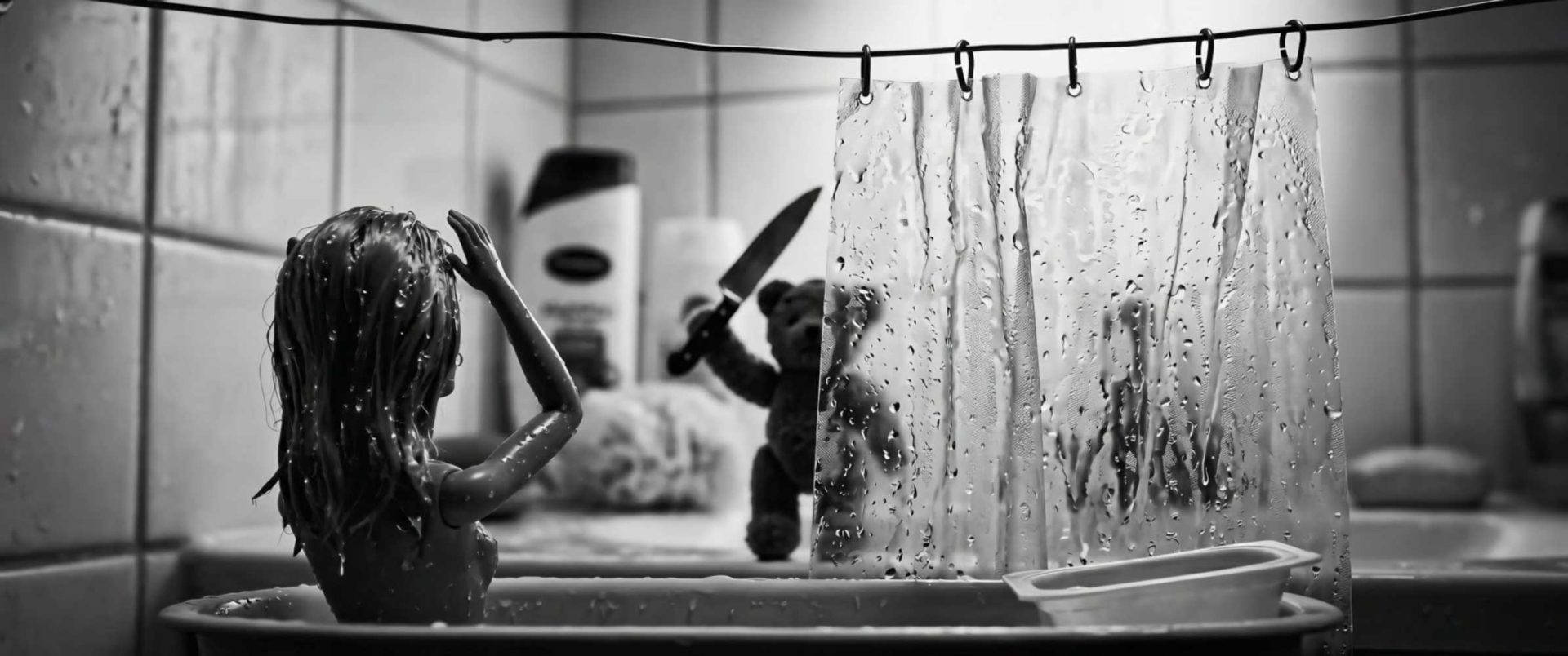 A doll sits in a bathtub with wet hair, while a small teddy bear holding a knife stands behind a clear shower curtain in a tiled bathroom—a cinephile’s nod to suspenseful horror, as if styled for the Young Director Awards. Frame from Stash Magazine article.