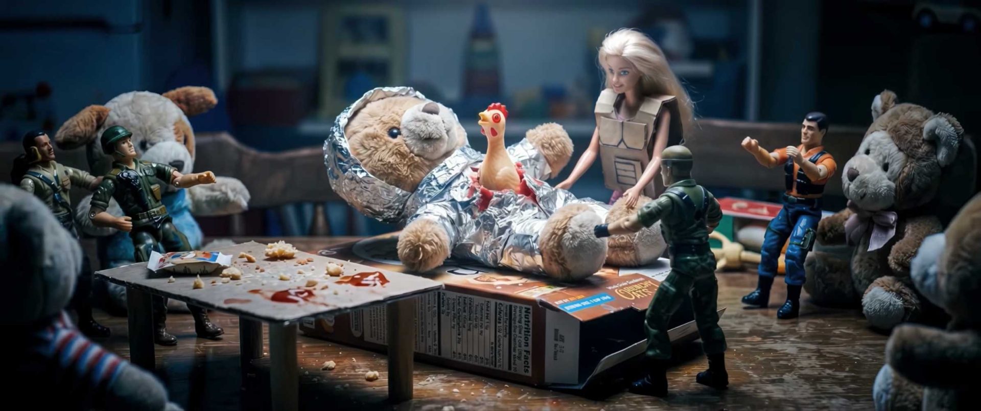 A group of toy soldiers and dolls, like cinephile judges at the Young Director Awards, stand around a teddy bear lying on a cereal box, wrapped in foil, with a rubber chicken on its chest. Crumbs and scattered objects cover the messy table. Frame from Stash Magazine article.