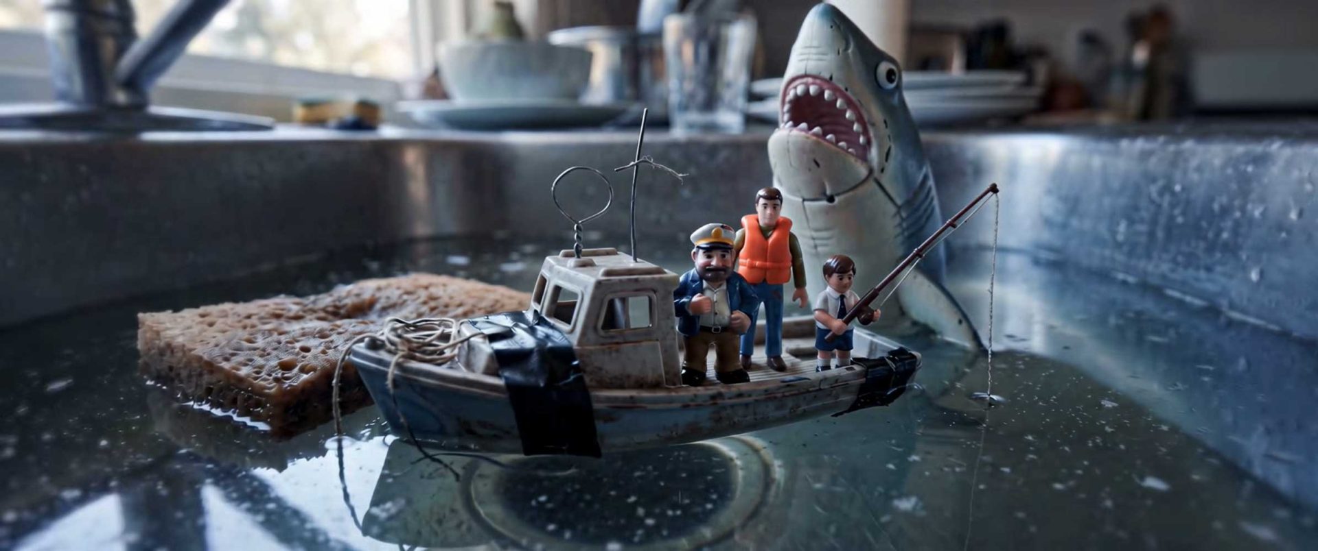 Three toy figures stand on a small boat floating in a kitchen sink filled with water, as a toy shark and sponge add playful drama—reminiscent of the creativity seen in the 2026 Young Director Awards opening titles by Wilders and Digital District. Frame from Stash Magazine article.