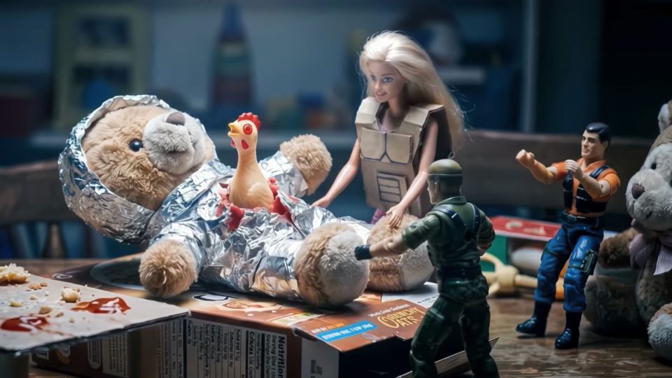 A teddy bear wrapped in foil lies on a table surrounded by toy soldiers, a Barbie doll, and a rubber chicken. The playful scene feels like an entry for the Young Director Awards, staged with quirky flair worthy of Digital District. Frame from Stash Magazine article.