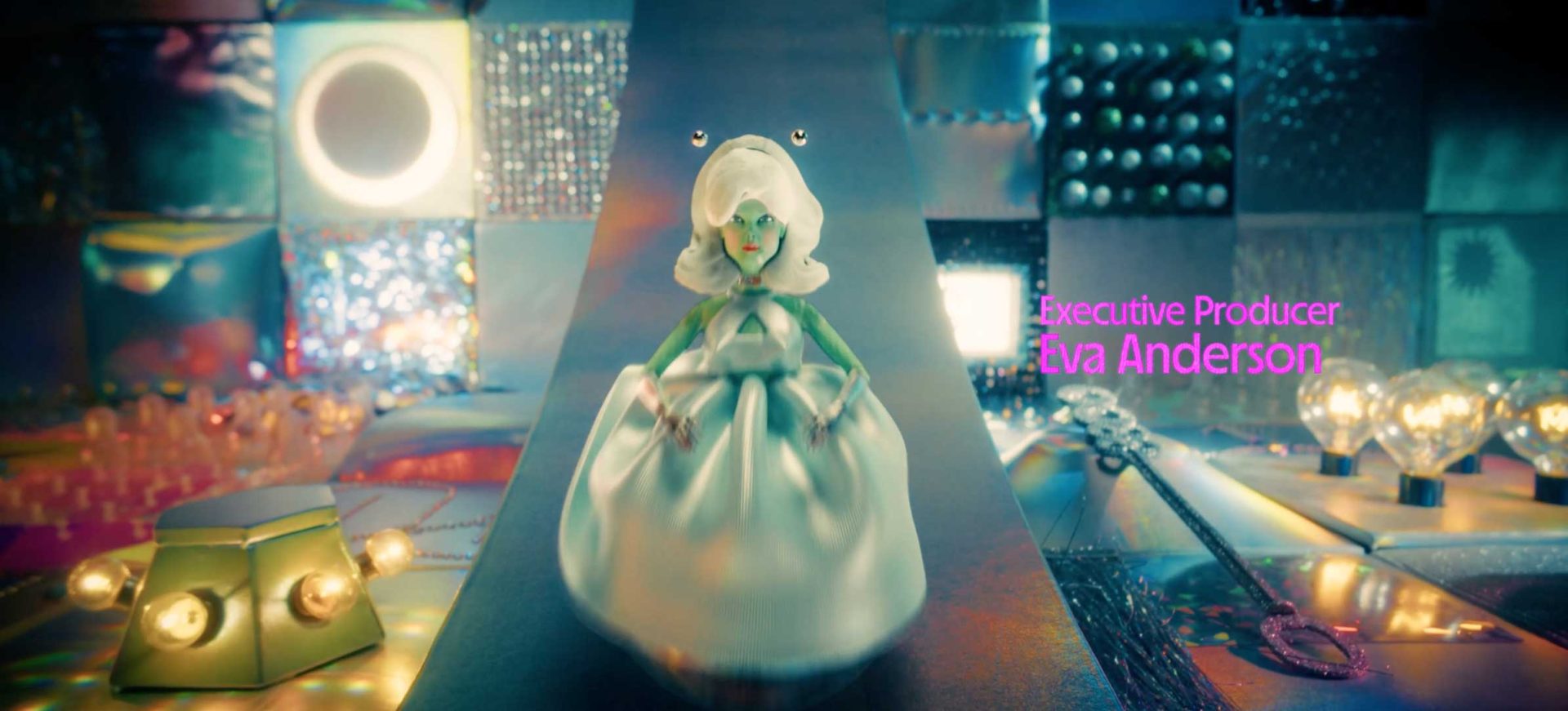 A doll-like figure in a white dress stands on a metallic, futuristic runway, surrounded by colorful, shiny panels and lightbulbs. Pink text reads Executive Producer Eva Anderson for Margo's Got Money Troubles on Apple TV from A24. Frame from Stash Magazine article.