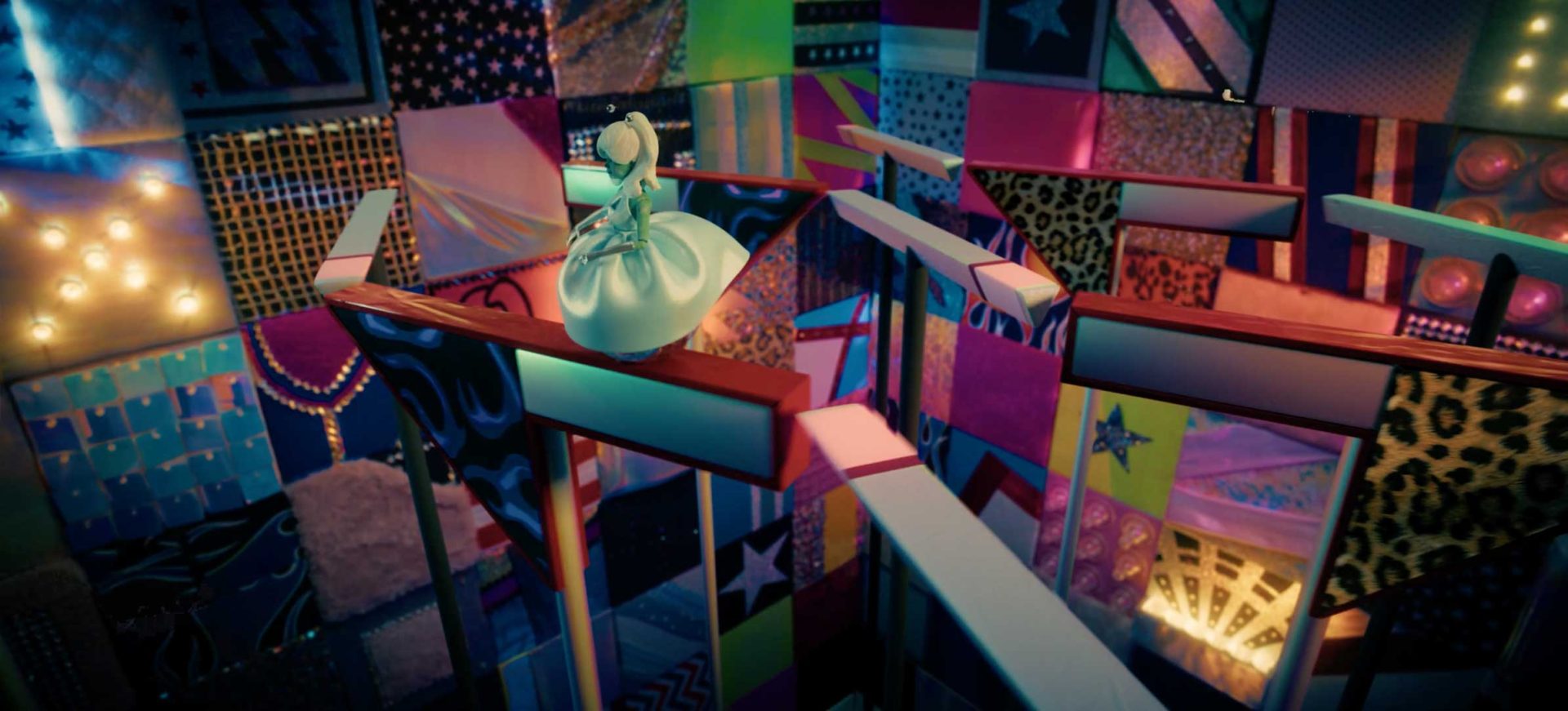 A doll in a white dress stands on a narrow, elevated path above a colorful, patchwork-patterned maze—evoking the surreal style of A24’s "Margo's Got Money Troubles" streaming now on Apple TV—with vibrant designs and varied textures on the walls. Frame from Stash Magazine article.