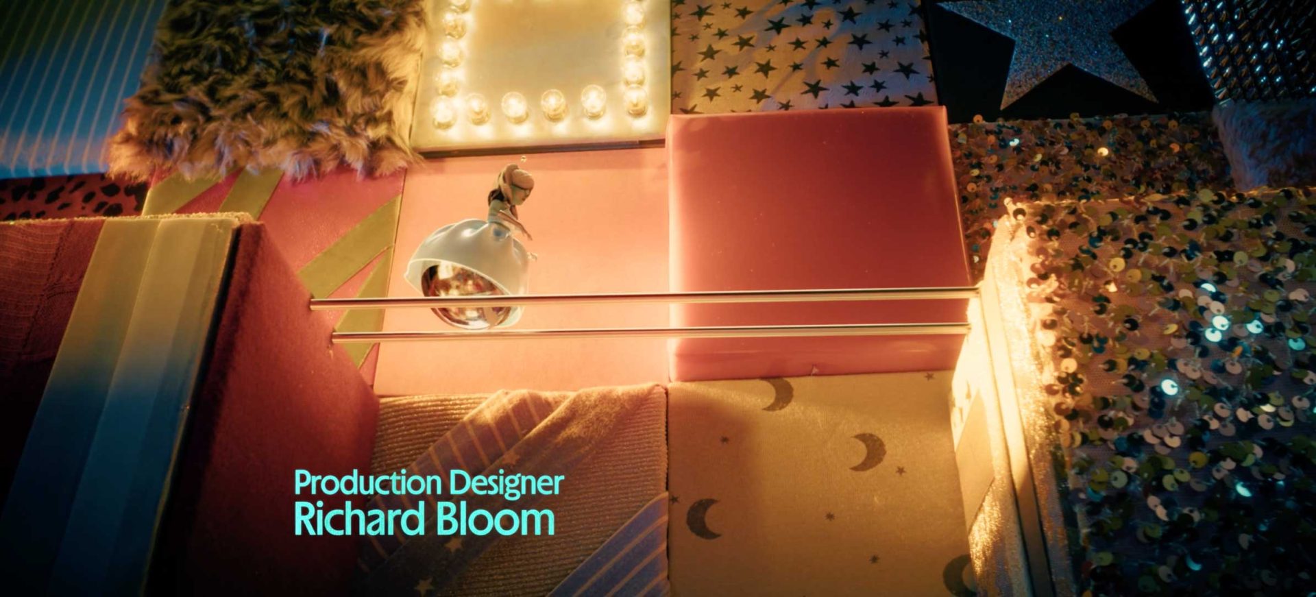 A wall decorated with various textured and patterned panels, including sequins, stars, and moons, plus a lamp fixture and neon light—reminiscent of Margo's Got Money Troubles Titles on Apple TV by Peter Anderson Studio. Text reads: "Production Designer Richard Bloom. Frame from Stash Magazine article.