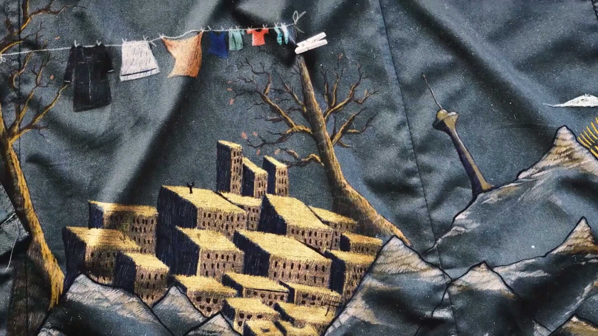 Embroidered art by Yegane Moghaddam shows yellow buildings on hills, a bare tree, a sword in the ground, and a clothesline with colorful garments—echoing the spirit of Our Uniform—set against a dark fabric background. Frame from Stash Magazine article.