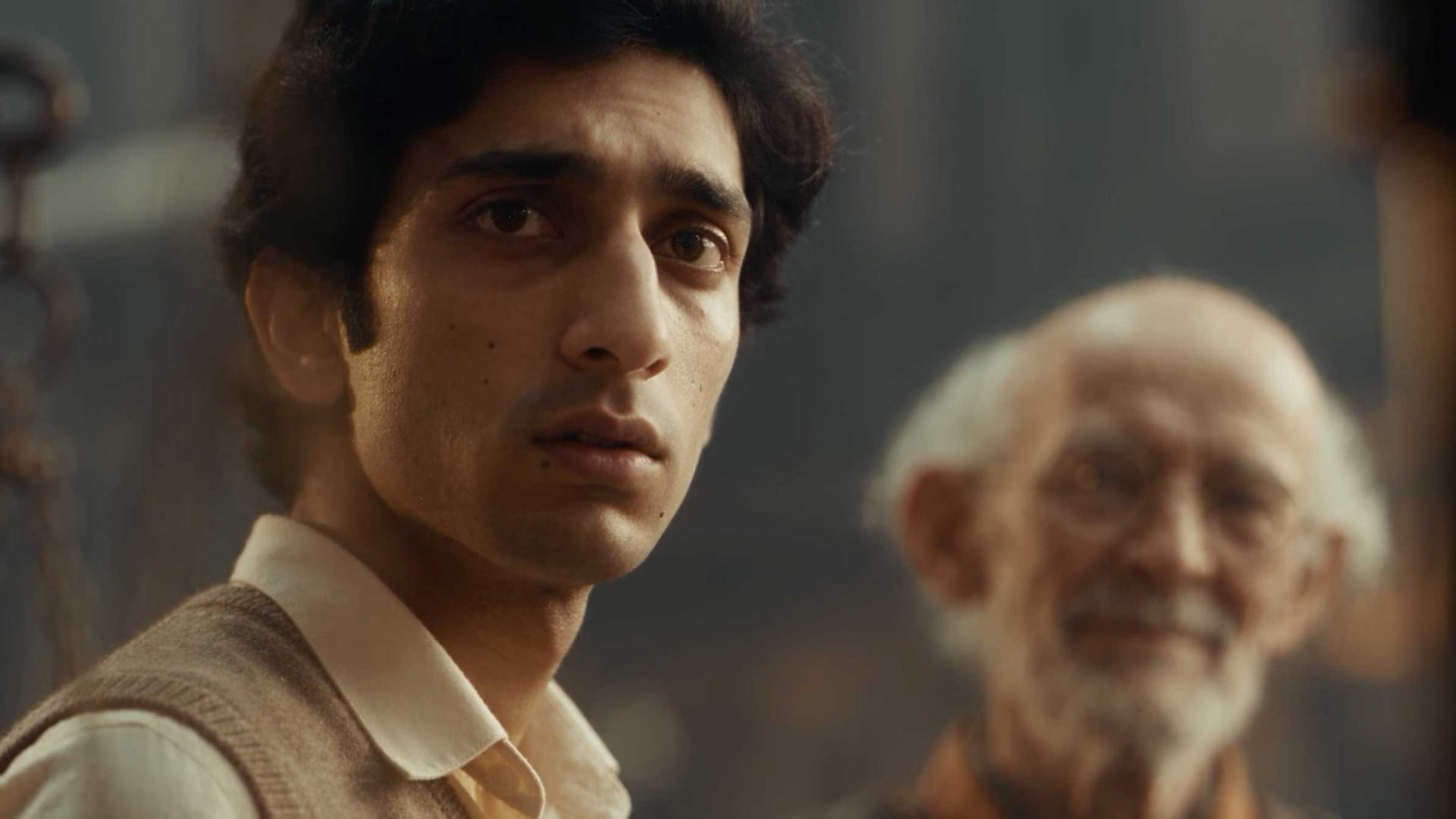 A young man with dark hair and a tan vest looks forward with a serious expression in this shot from the short film Query, while an older man with white hair and glasses stands blurred in the background. Frame from Stash Magazine article.