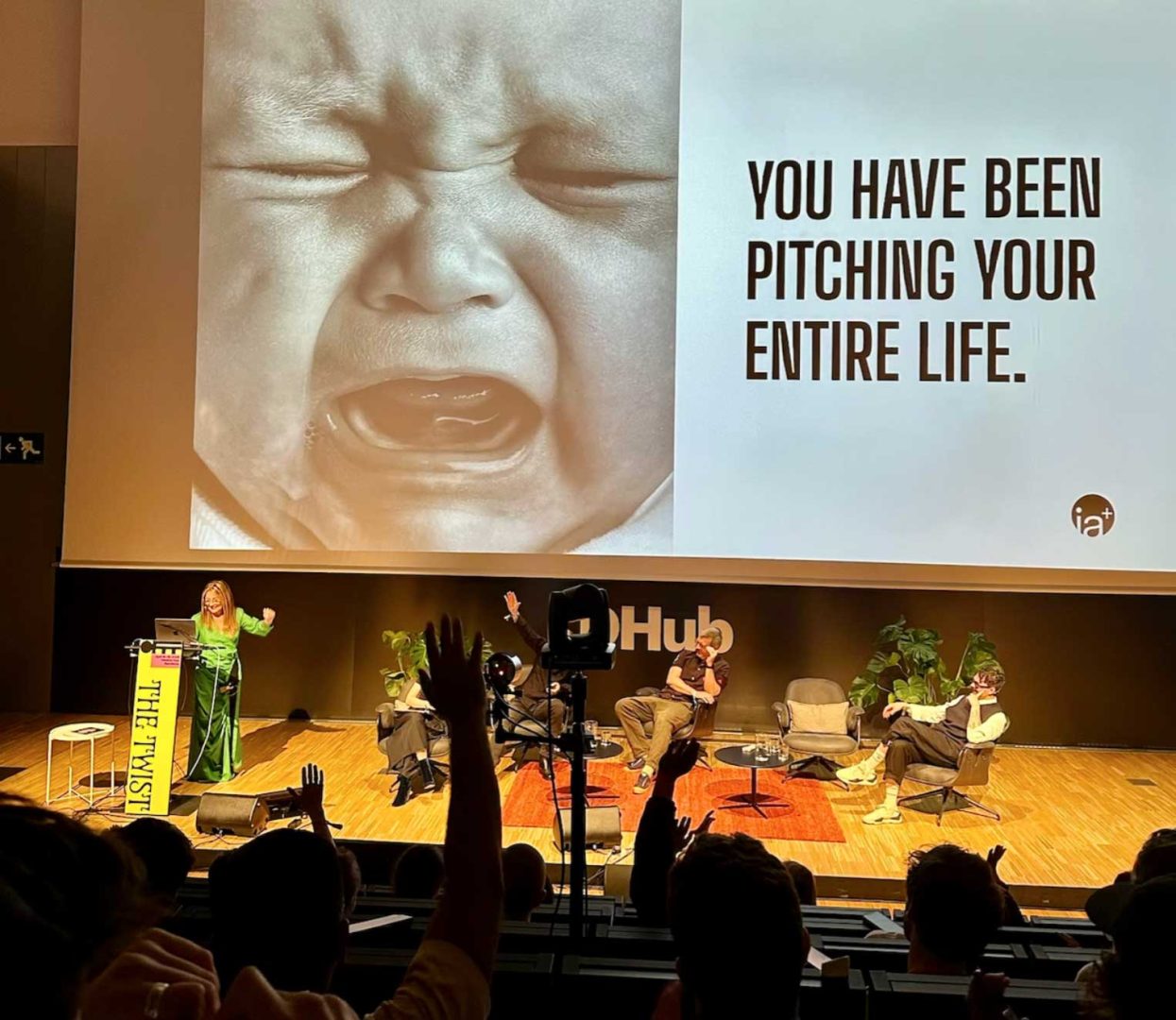 A woman speaks on stage at a conference beside a screen displaying a crying baby and the text YOU HAVE BEEN PITCHING YOUR ENTIRE LIFE, referencing Life's a Pitch: 9 Pitching Rules to Stop Giving Your Value Away. Audience members raise their hands; three panelists are seated. Frame from Stash Magazine article.