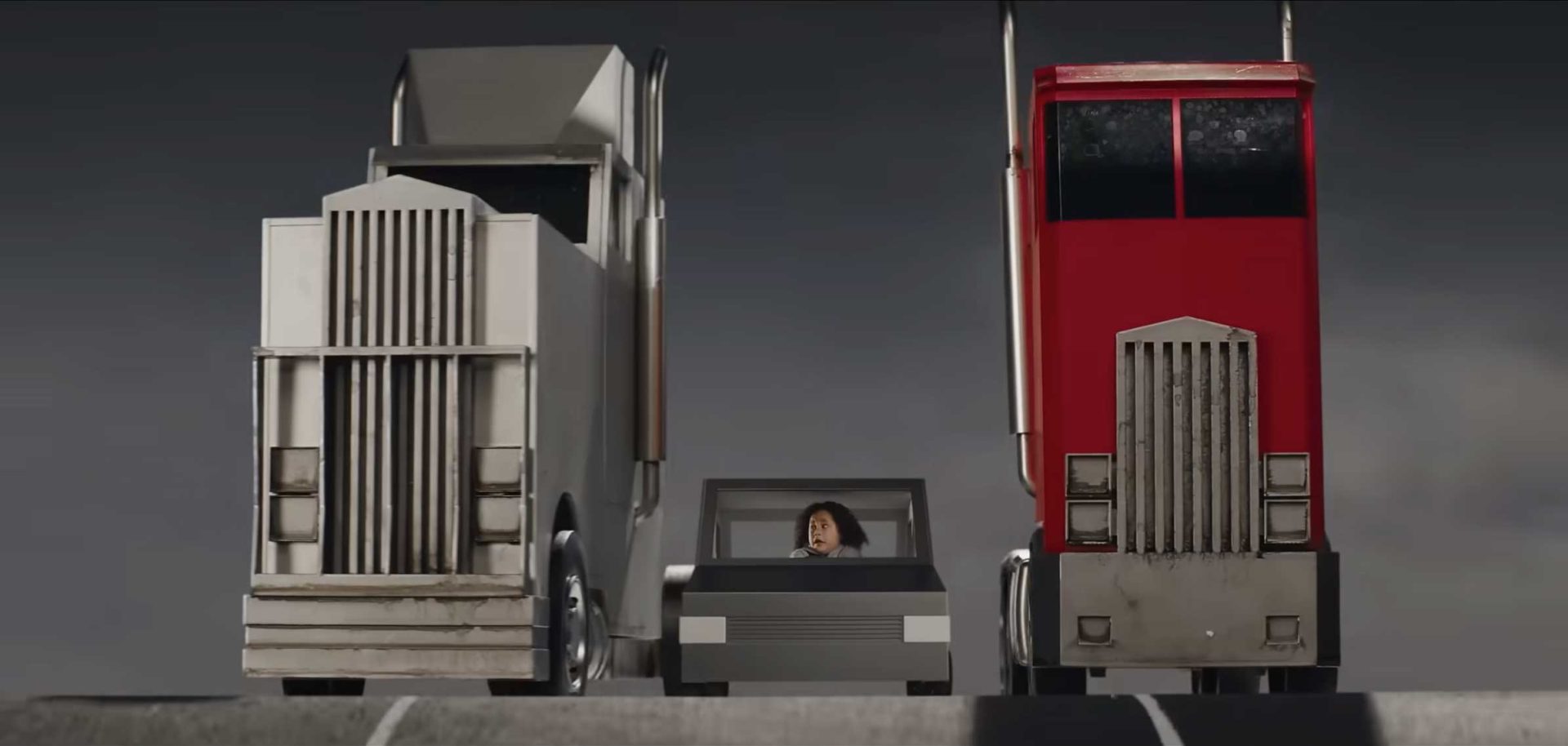 A person in a small gray car, perhaps contemplating whether to call AA Insurance, looks up nervously while driving between a large white truck on the left and a massive red truck on the right under a gray sky—a scene evocative of Joel Kefali’s cinematic style. Frame from Stash Magazine article.