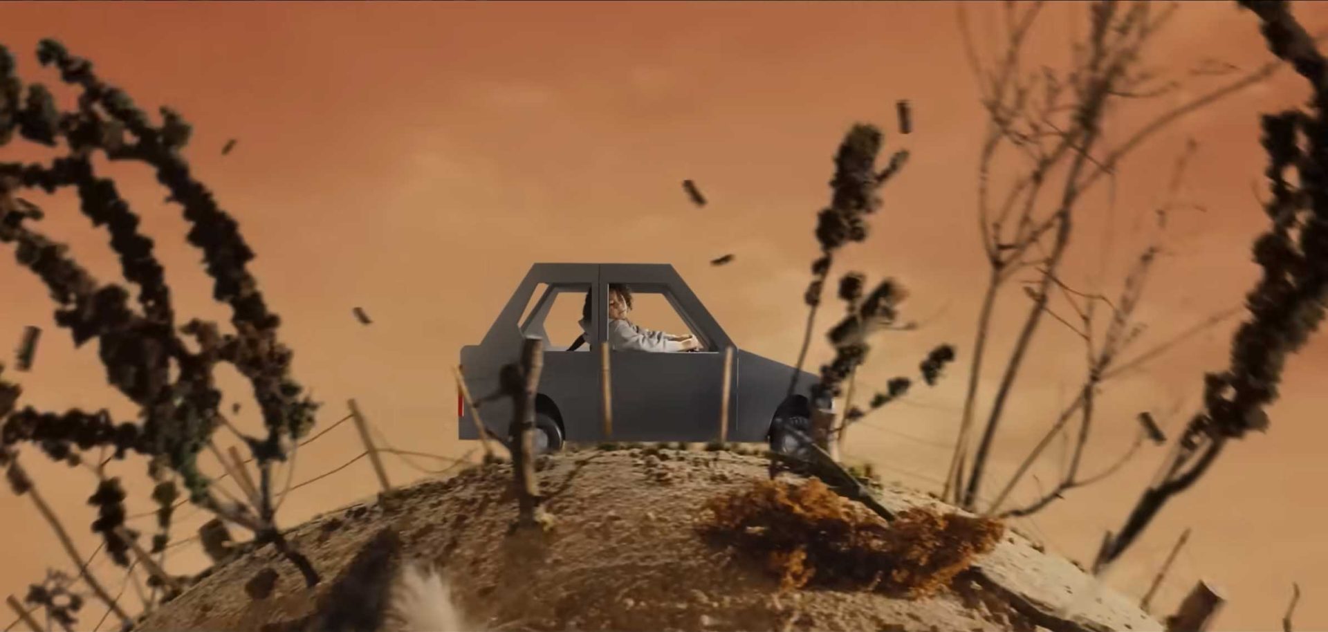 A person sits in a gray, boxy car atop a small hill with sparse, dry plants, set against an orange sky. The scene has a surreal, almost animated appearance, reminiscent of Joel Kefali’s unique visual style. Frame from Stash Magazine article.