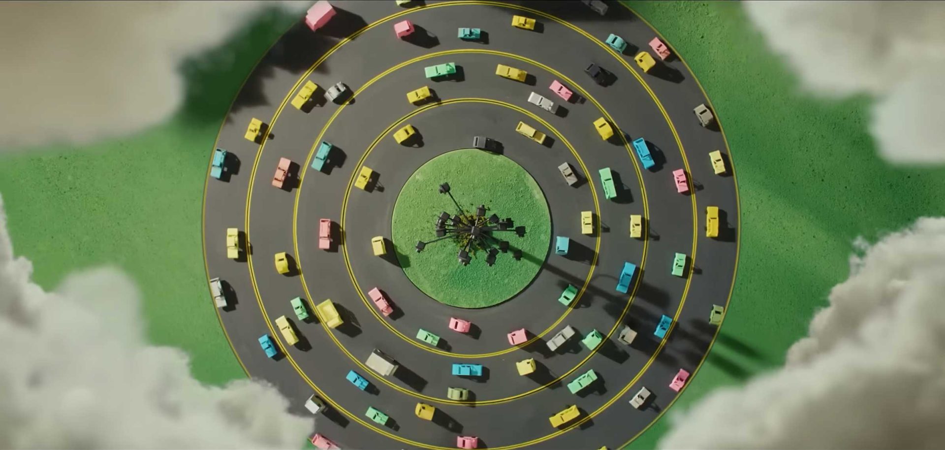 Aerial view of colorful toy cars driving in three concentric circular roads surrounding a small green park with trees in the center, with clouds partially surrounding the scene—an imaginative setup reminiscent of the playful visuals from the AA Insurance Born Ready Ad by Joel Kefali and Good Oil. Frame from Stash Magazine article.