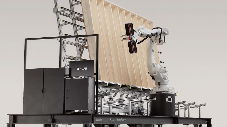 A robotic arm assembles a large wooden panel on a metal frame, surrounded by machinery labeled AUAR. The setup, part of the innovative Robot MicroFactories developed by AUAR and XO3D, appears to be an advanced automated construction or manufacturing workstation. Frame from Stash Magazine article.