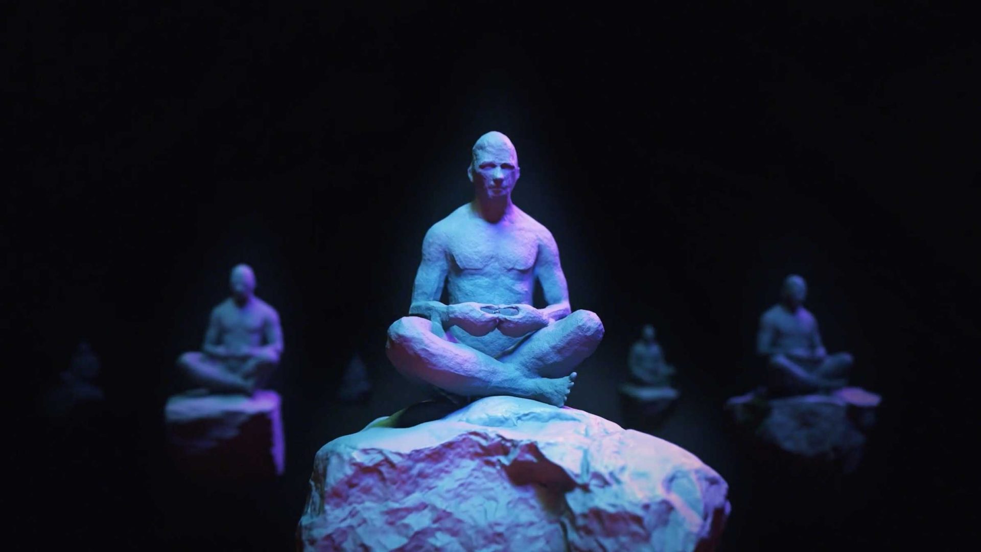 A blue-toned statue of a person sits cross-legged on a floating rock against a dark background, reminiscent of João Vaz Oliveira’s style. Similar statues meditate on distant rocks, illuminated by soft purple and blue light. Frame from Stash Magazine article.