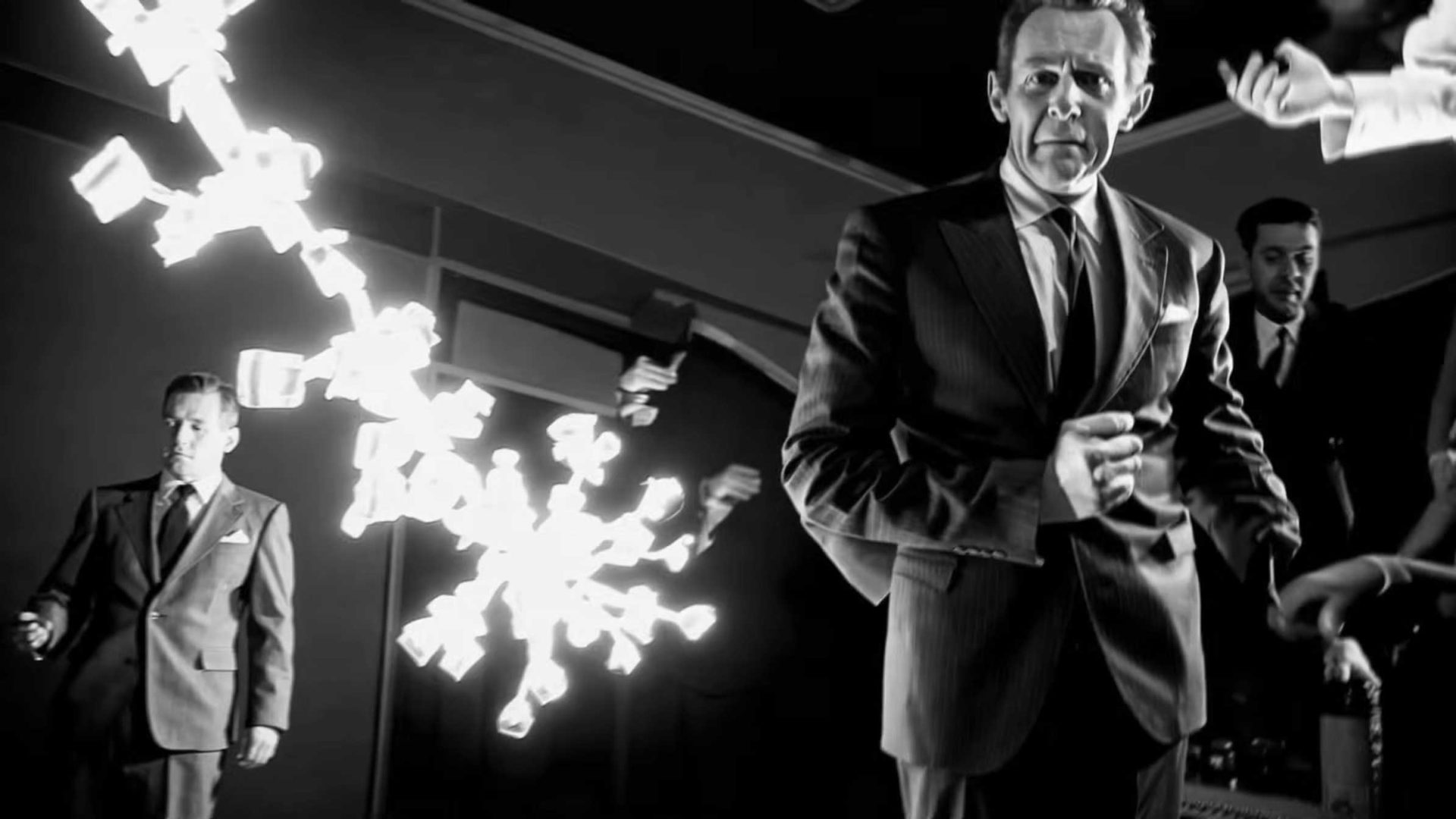Men in suits stand in a room as bills fly through the air; one man in front looks serious while others react to the chaos, all rendered in striking black and white reminiscent of Alessandro Bavari’s surreal style, as if illuminated by solid light from space. Frame from Stash Magazine article.