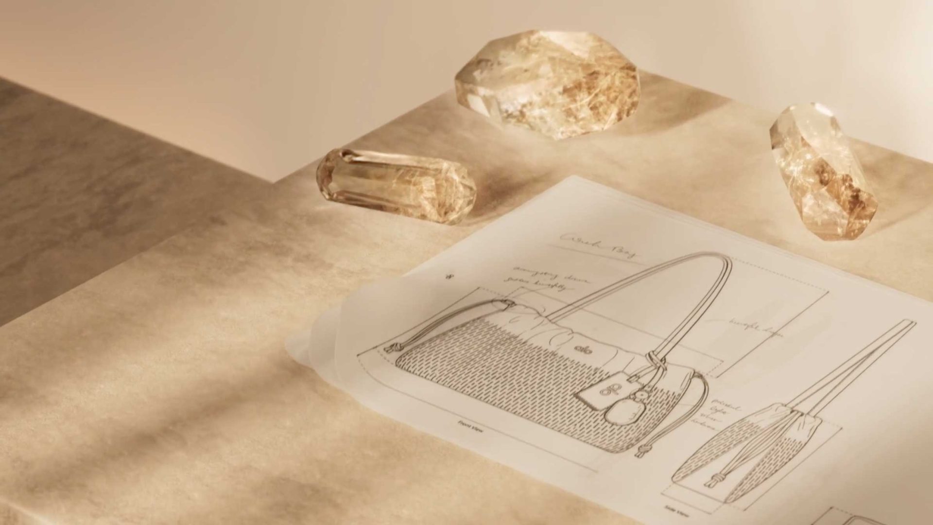 A technical sketch of an Alo handbag with handwritten annotations, resting on a stone surface alongside three large crystals, evoking a sense of quiet luxury. Frame from Stash Magazine article.