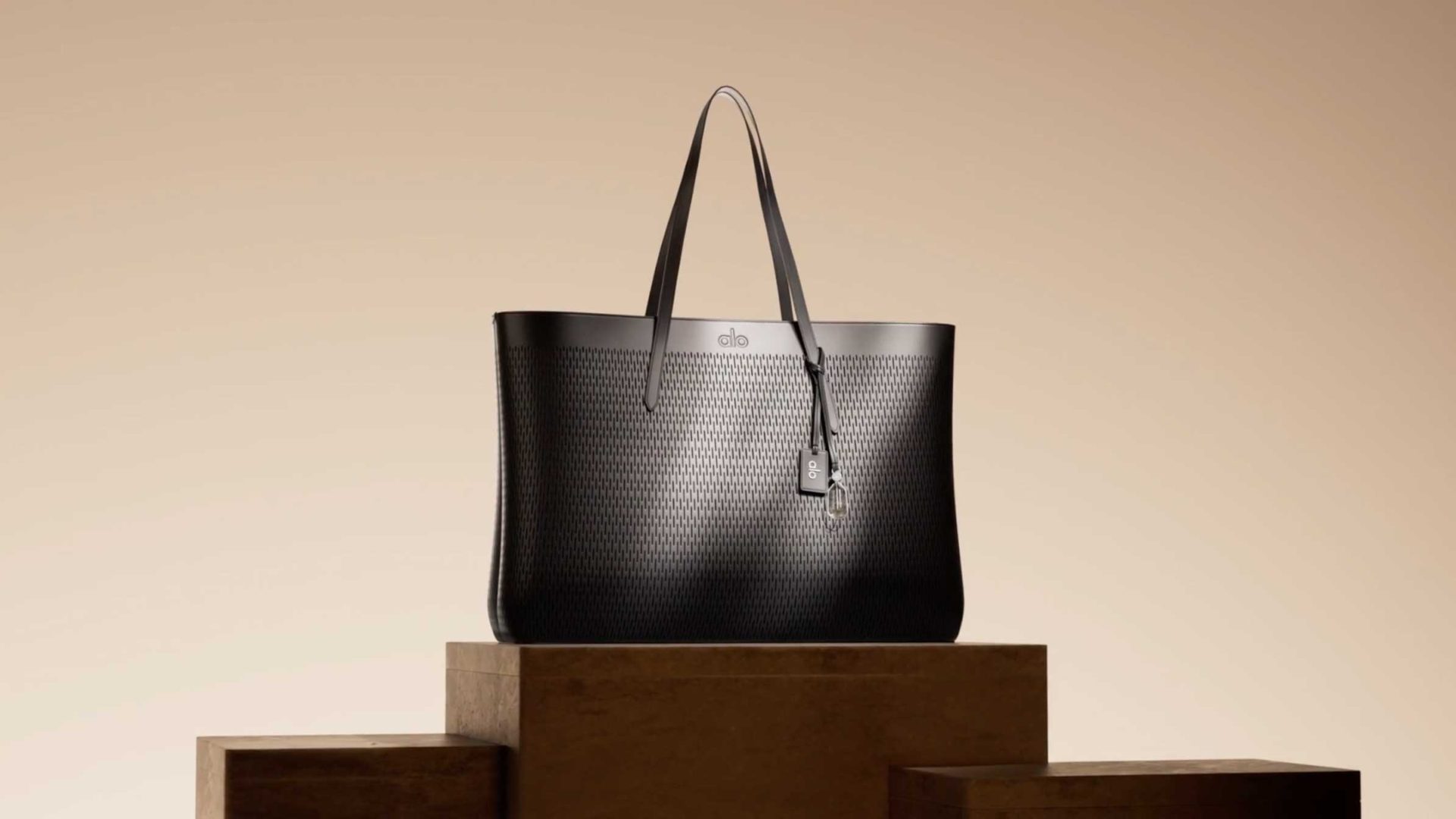 Black Tendril Intro tote bag with a perforated pattern and an Alo logo tag, sitting on a tiered wooden platform in a quiet luxury style. Frame from Stash Magazine article.