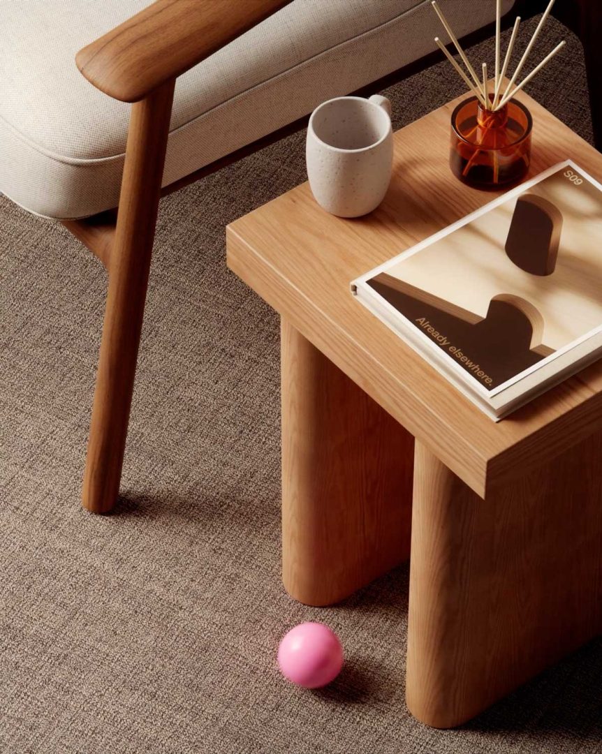 A modern wooden side table styled with a mug, brown glass diffuser, and art book sits beside a beige cushioned chair; a pink ball inspired by Andreas Wannerstedt’s Soft Logic designs rests on the carpeted floor below. Frame from Stash Magazine article.
