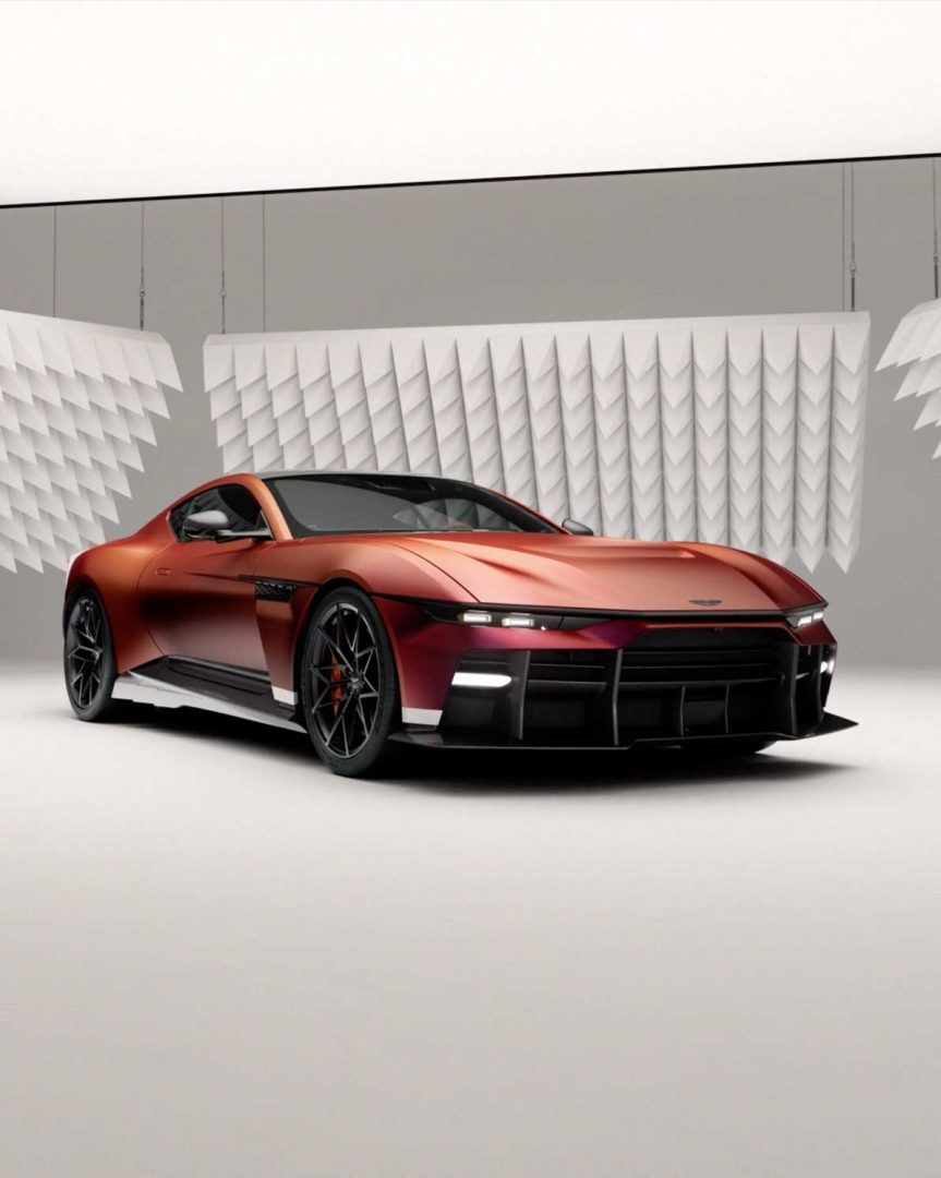 A sleek, metallic red Aston Martin sports car with black accents is displayed in a modern studio with geometric white panel decor hanging behind it. Frame from Stash Magazine article.