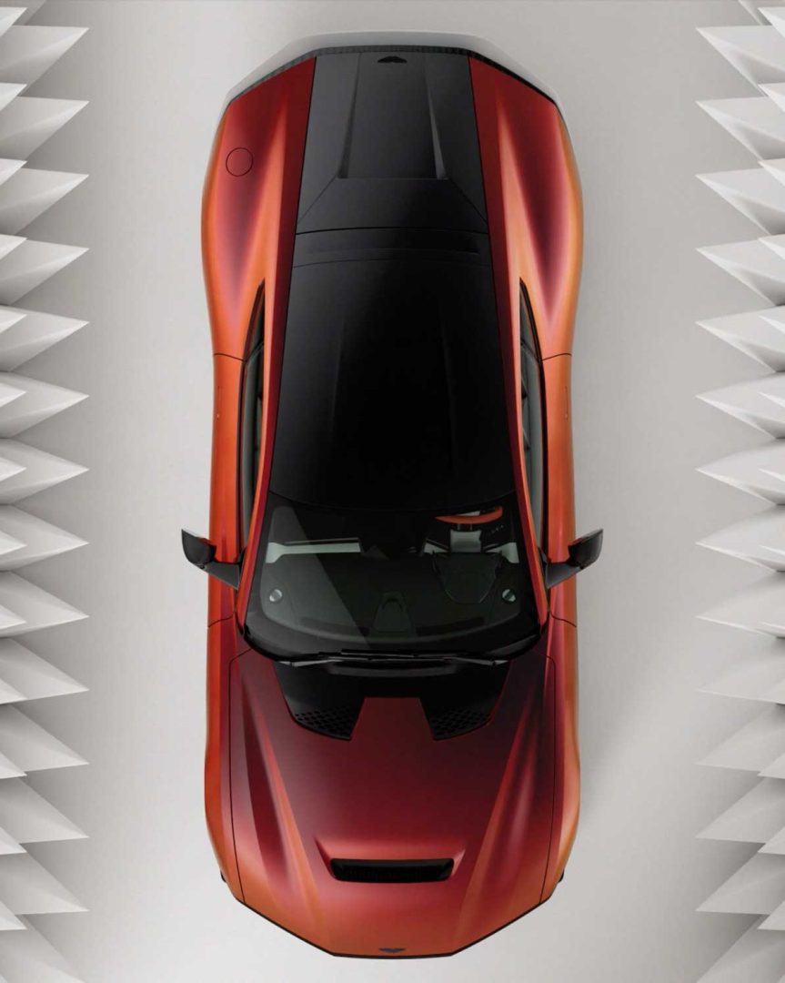 Top-down view of a shiny orange Valen Supercar with black accents, positioned between patterned white, triangular wall panels on both sides. Frame from Stash Magazine article.