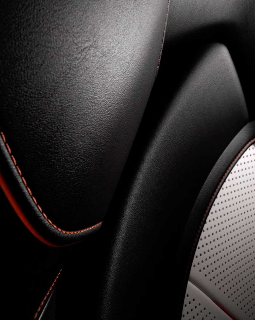 Close-up of a car seat with black leather upholstery, orange stitching, and a section of perforated white material, highlighting texture and detail in the design—reflecting the signature craftsmanship found in an Aston Martin interior. Frame from Stash Magazine article.