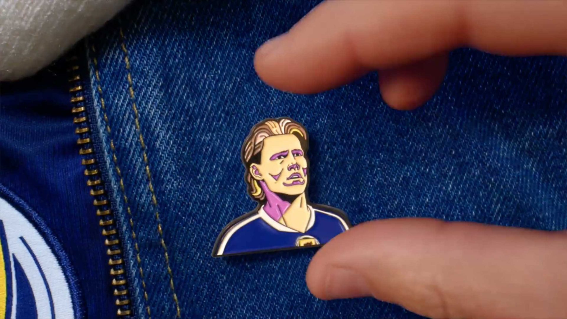 A hand holds an enamel pin depicting a stylized portrait, inspired by Nicos Livesey’s animation for the BBC World Cup, featuring a person with shoulder-length hair and a dark shirt, pinned to blue denim fabric next to a brass zipper. Frame from Stash Magazine article.