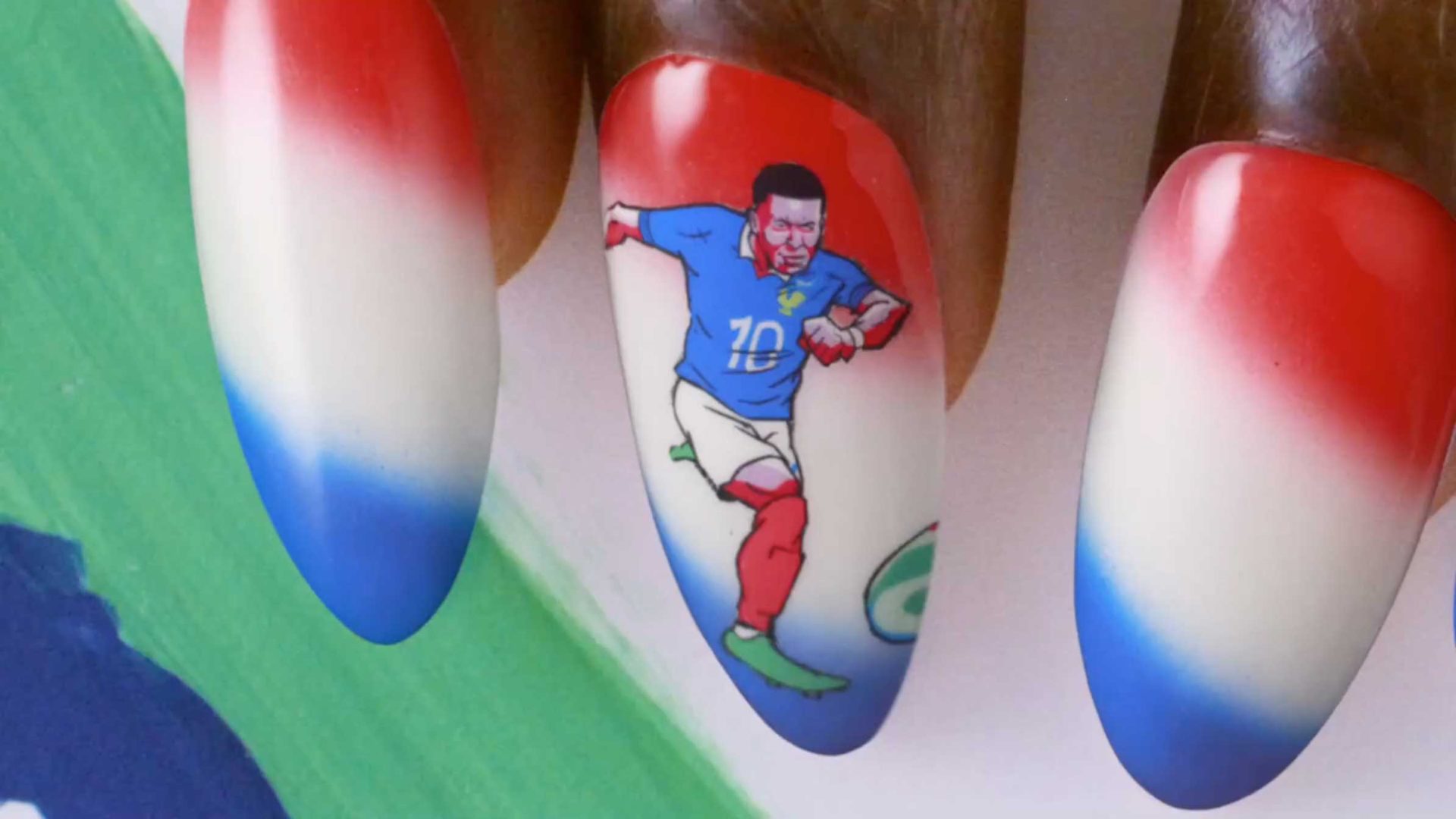 Close-up of fingernails painted in red, white, and blue; one nail features detailed nail art inspired by Nicos Livesey’s BBC World Cup animation, showing a soccer player in a blue jersey kicking a ball. Frame from Stash Magazine article.