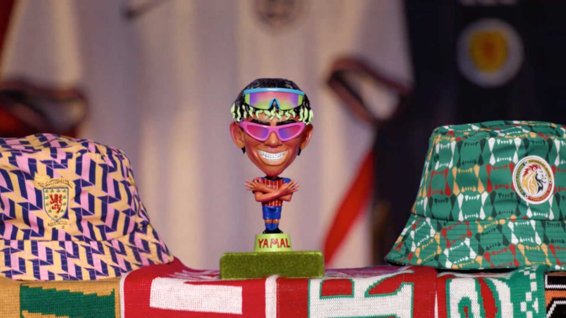 A bobblehead with sunglasses and a colorful headband stands between two patterned bucket hats on a table draped with vibrant scarves. The base reads YAMAL. Animation-inspired sports jerseys, reminiscent of Nicos Livesey’s BBC World Cup style, hang in the background. Frame from Stash Magazine article.