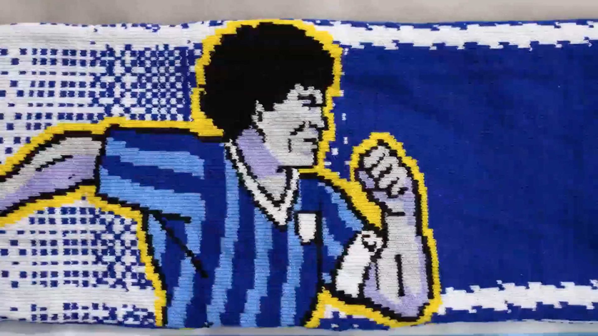 A woven textile depicts a stylized soccer player in a blue and black striped jersey, running with a clenched fist, inspired by the BBC World Cup 2026 Coverage animated trailer by Nicos Livesey and Blinkink. Blue background with yellow accents adds vibrancy. Frame from Stash Magazine article.