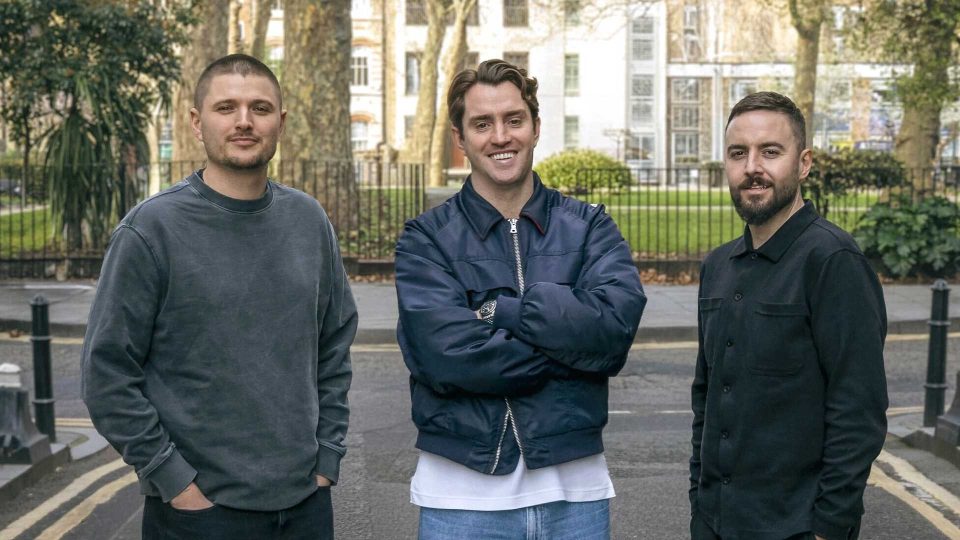 BRASH launches in London with co-founders Alasdair Patrick, Tom Clapp and Matt Fuller. Three men stand on a street in London, in front of trees and a park fence. The man in the center smiles with arms crossed, while the others stand casually on either side, all dressed in casual clothes.