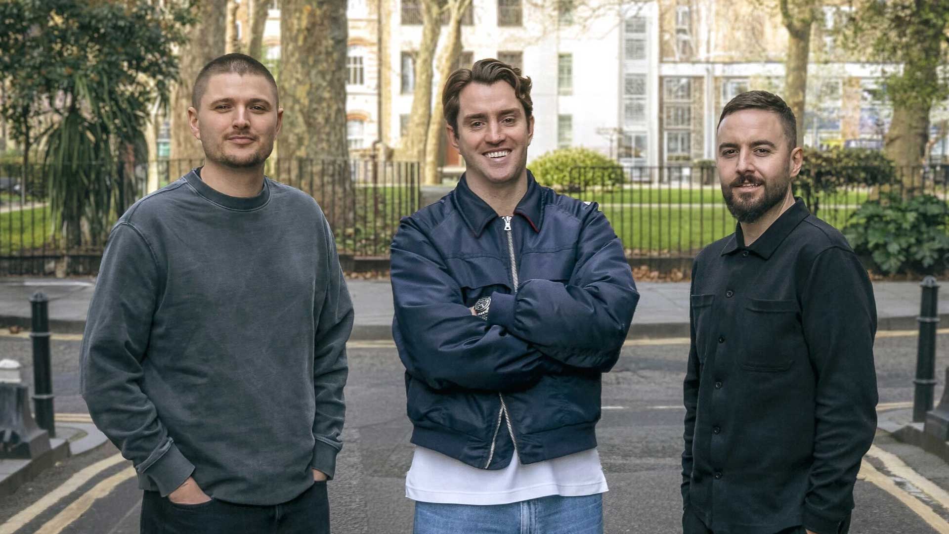 BRASH launches in London with co-founders Alasdair Patrick, Tom Clapp and Matt Fuller. Three men stand on a street in London, in front of trees and a park fence. The man in the center smiles with arms crossed, while the others stand casually on either side, all dressed in casual clothes.