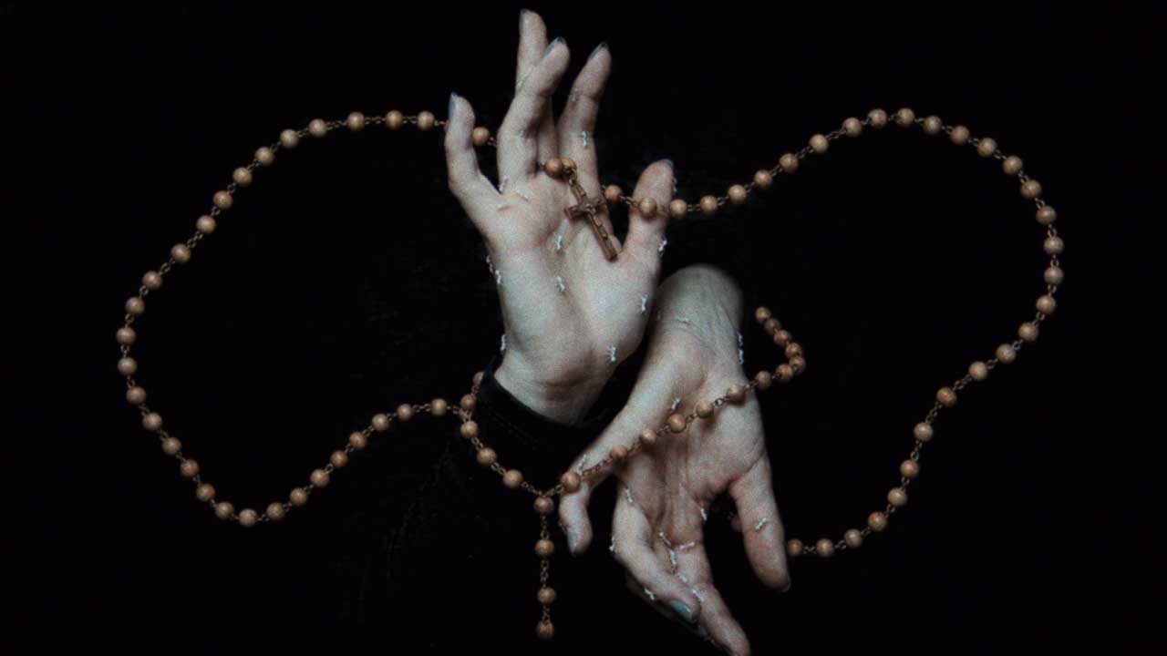 Two pale hands intertwined against a dark background, with a wooden rosary wrapped around the fingers, evoking the tense atmosphere of Sijin Liu’s "Ici (Remix)" amidst drifting echoes like bai yun—white clouds. Frame from Stash Magazine article.