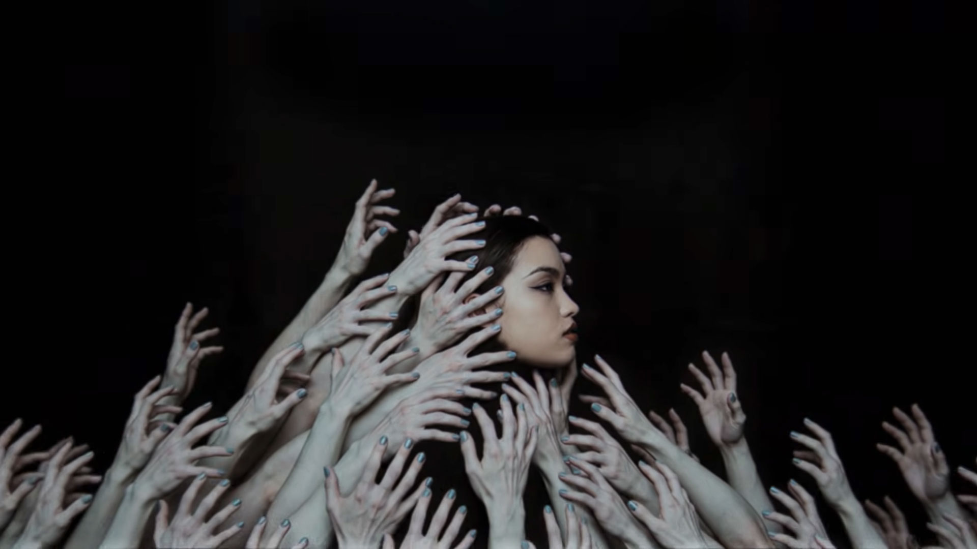 A woman with a calm expression is surrounded by numerous pale hands reaching toward her from all directions against a dark background, evoking the haunting atmosphere of Sijin Liu's artwork or the mood in Bai Yun’s Ici (Remix). Frame from Stash Magazine article.