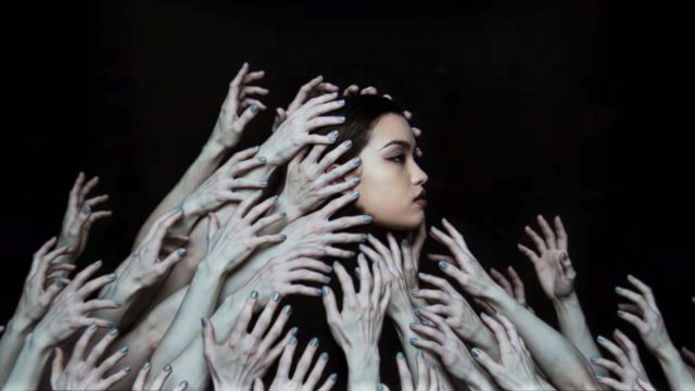 A woman with long dark hair looks to the side, surrounded by numerous pale hands reaching toward her against a black background—echoing the sense of overwhelm found in the Bai yun Ici remix music video by Sijin Liu. Frame from Stash Magazine article.