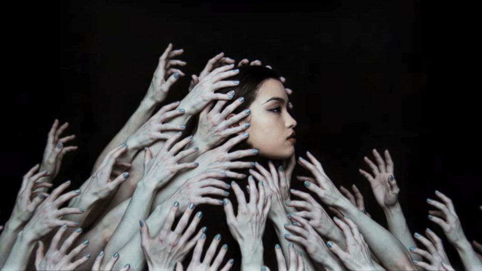 A woman with long dark hair looks to the side, surrounded by numerous pale hands reaching toward her against a black background—echoing the sense of overwhelm found in the Bai yun Ici remix music video by Sijin Liu. Frame from Stash Magazine article.