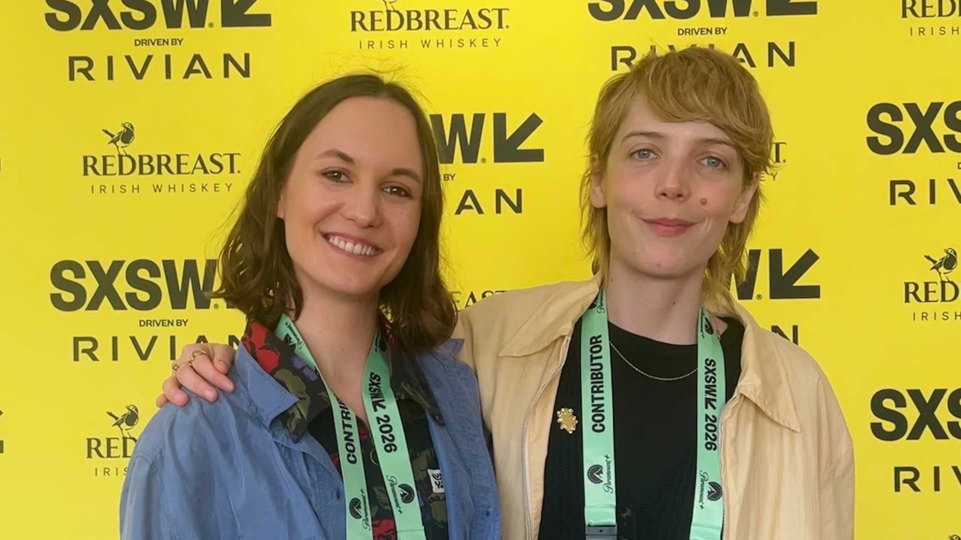 Two women stand smiling together in front of a bright yellow SXSW photo backdrop featuring sponsor logos. They both wear light jackets and event lanyards, and one has her arm around the other. Frame from Stash Magazine article.