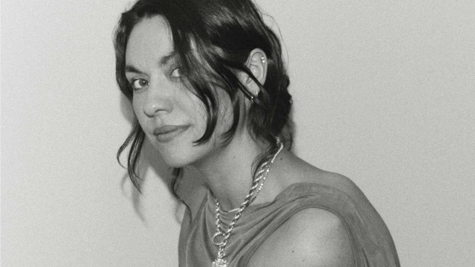 Black and white photo of a person with wavy dark hair, layered chain necklaces, and a sleeveless top, looking over their shoulder—capturing the bold style that caught attention when Biscuit Filmworks signs Phoebe Arnstein for commercial representation. Frame from Stash Magazine article.