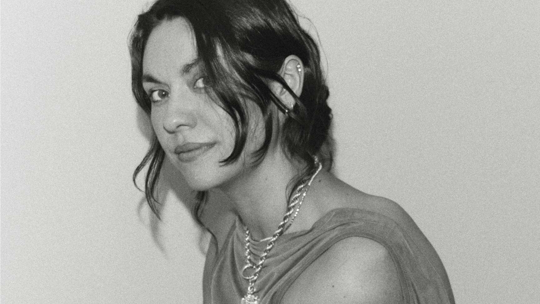 Black and white photo of a person with wavy dark hair, layered chain necklaces, and a sleeveless top, looking over their shoulder—capturing the bold style that caught attention when Biscuit Filmworks signs Phoebe Arnstein for commercial representation. Frame from Stash Magazine article.
