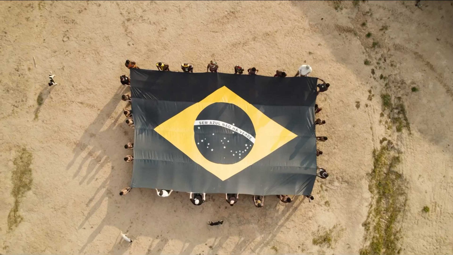 A group of people stand in a circle on sandy ground, holding a large Brazilian flag viewed from above. Their shadows surround the flag as they unite to support SOS Oceano and raise awareness for ocean protection. Frame from Stash Magazine article.