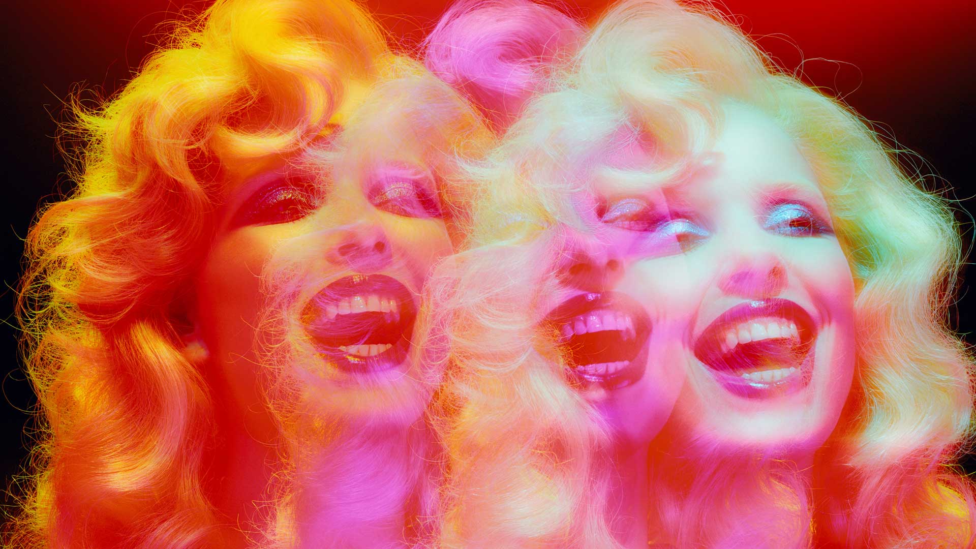 A vibrant, psychedelic image reminiscent of Miles Aldridge’s bold style: a person with wavy blond hair and bright makeup, shown with a multiple exposure effect creating three overlapping, colorful faces, all laughing joyfully under vivid red and pink lighting. Frame from Stash Magazine article.