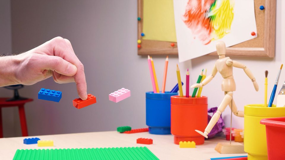 A hand drops colorful toy bricks onto a desk with art supplies, colored pencils, a wooden mannequin, and a child’s painting on the wall in the background—reminiscent of the playful creativity found in stop-motion director Kevin Parry’s work. Frame from Stash Magazine article.