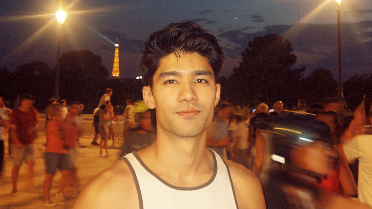 A young man in a white tank top stands outdoors at night, with blurred people around him and the Eiffel Tower illuminated in the background—evoking a cinematic atmosphere reminiscent of Blinkink Signs Australian Director Radheya Jang for Worldwide Representation. Frame from Stash Magazine article.