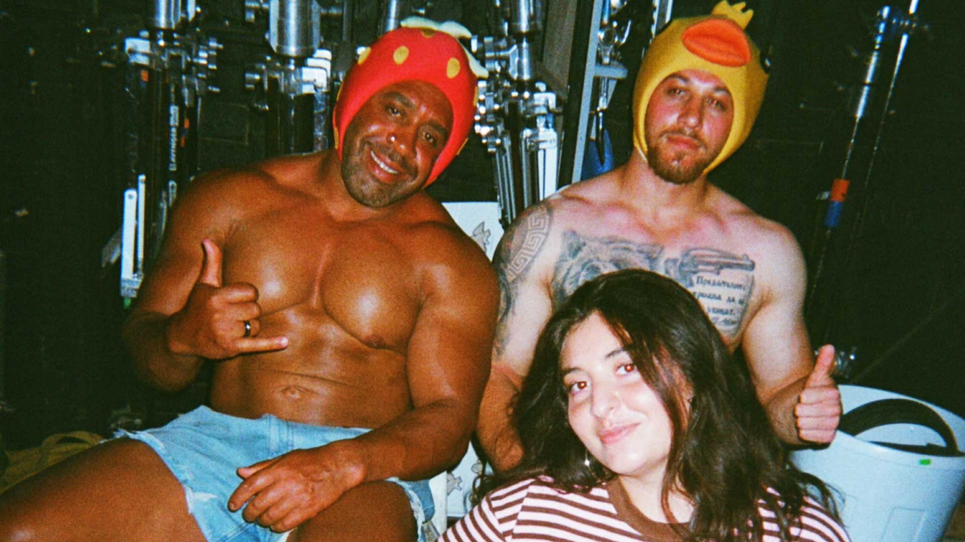 Three people pose together. Two shirtless men in funny hats (a ladybug and a duck) sit with a smiling woman in a striped shirt in front of them. The background has metal equipment and dim lighting, capturing a quirky scene that aligns perfectly with Blinkink to represent director Léa Esmaili in the US market. Frame from Stash Magazine article.