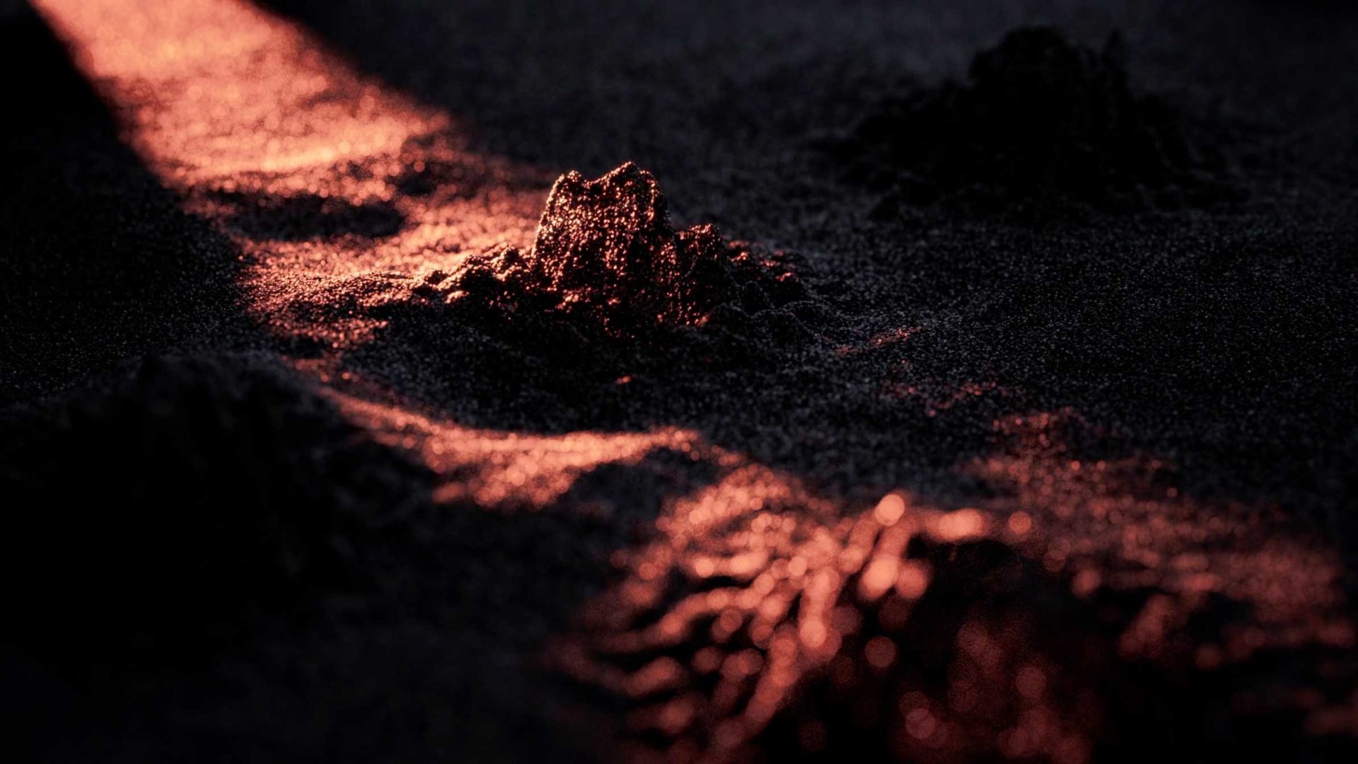Mounds of dark sand are softly illuminated by streaks of warm, orange light, casting dramatic shadows and highlighting the rough, textured surface in this Bloq.it Smart Lockers product film by Depth of Field. Frame from Stash Magazine article.