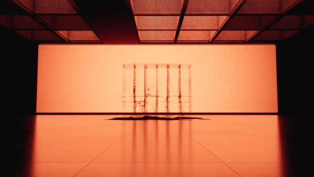 Minimalist scene with an orange glow, featuring a transparent, cage-like structure at the center, reminiscent of a Bloq.it Smart Lockers product film by Depth of Field. The ceiling’s grid pattern and long shadows enhance the modern, polished space. Frame from Stash Magazine article.