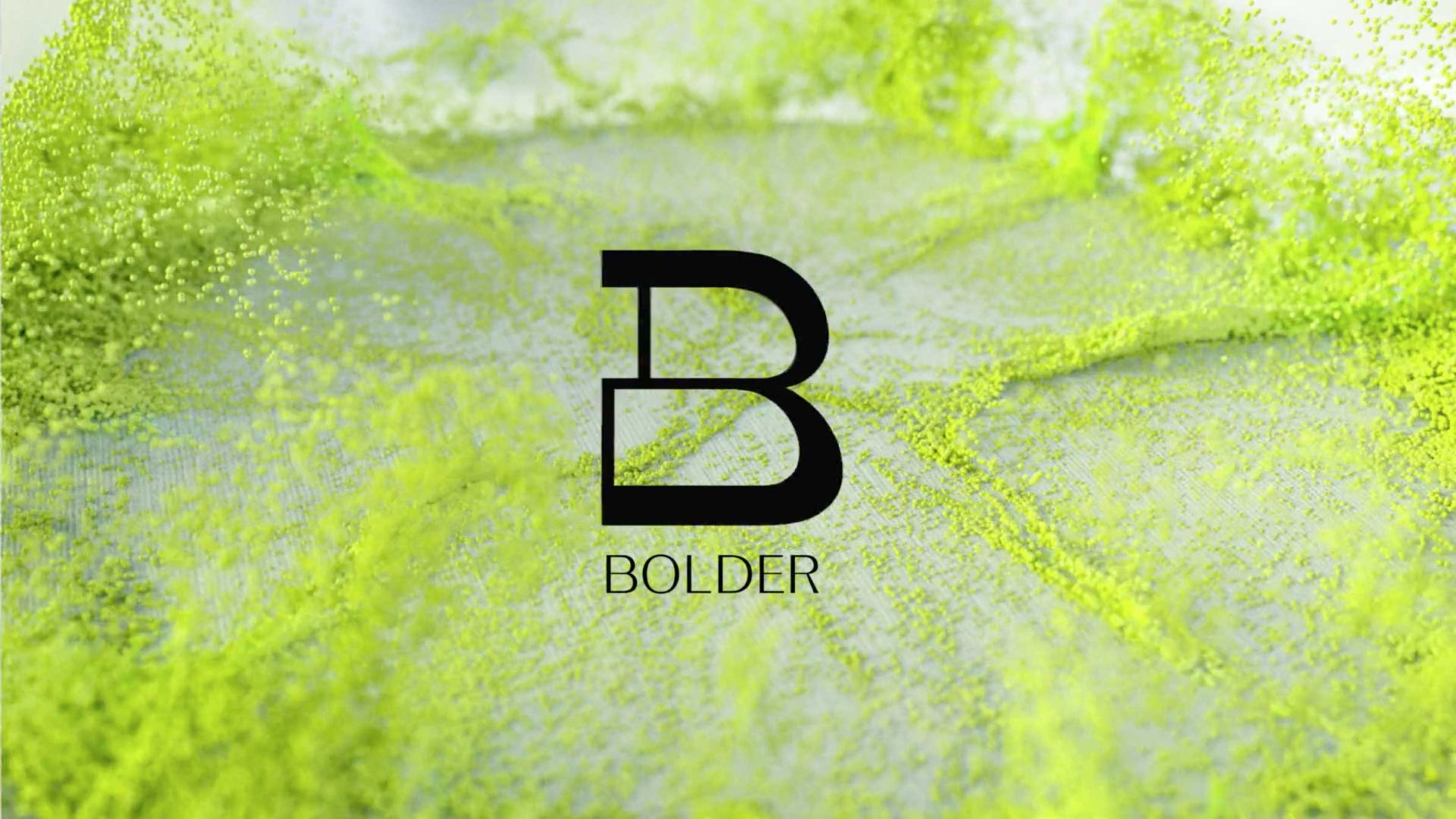 A large black letter B with the word BOLDER beneath it appears at the center of a background featuring vibrant splashes and streaks of bright green liquid in motion, creating a dynamic and energetic design. Frame from Stash Magazine article.