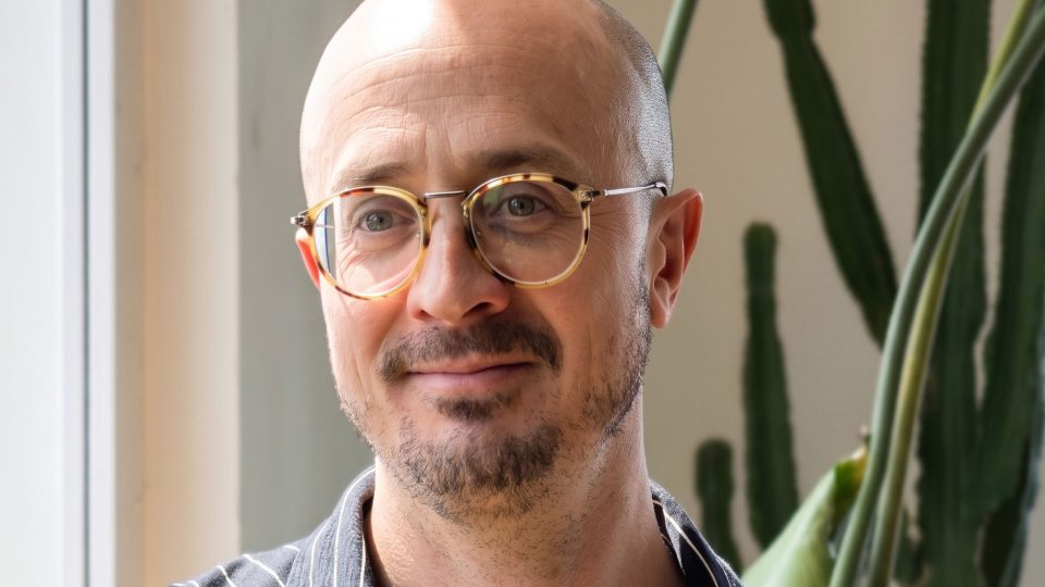 A person with glasses and a trimmed beard smiles slightly while standing near a window, with a green cactus plant in the background, reflecting the collaborative spirit behind Builders Club Appointments and Matt Shannon’s new role as Executive Producer Partnerships. Frame from Stash Magazine article.