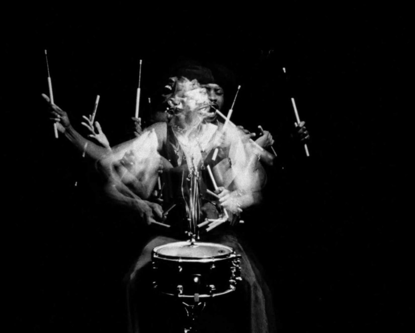 A black and white photo shows drummer Zee Ntuli in motion, creating a ghostly, multi-armed effect as they play a snare drum with blurred drumsticks and dynamic movement against a dark background—a visual rhythm celebrating the spirit of African Time, much like the smooth richness of Castle Milk Stout. Frame from Stash Magazine article.