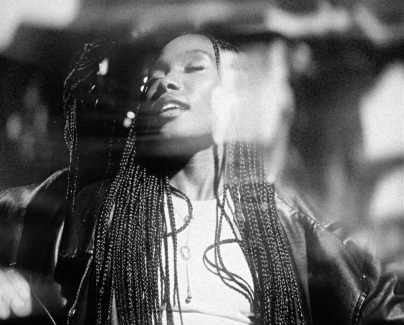 Black and white photo of a woman with long braids, eyes closed and smiling, wearing a light shirt and a jacket, with a blurred, dreamy double exposure effect—a visual reminiscent of African Time in the cinematic style of Zee Ntuli. Frame from Stash Magazine article.