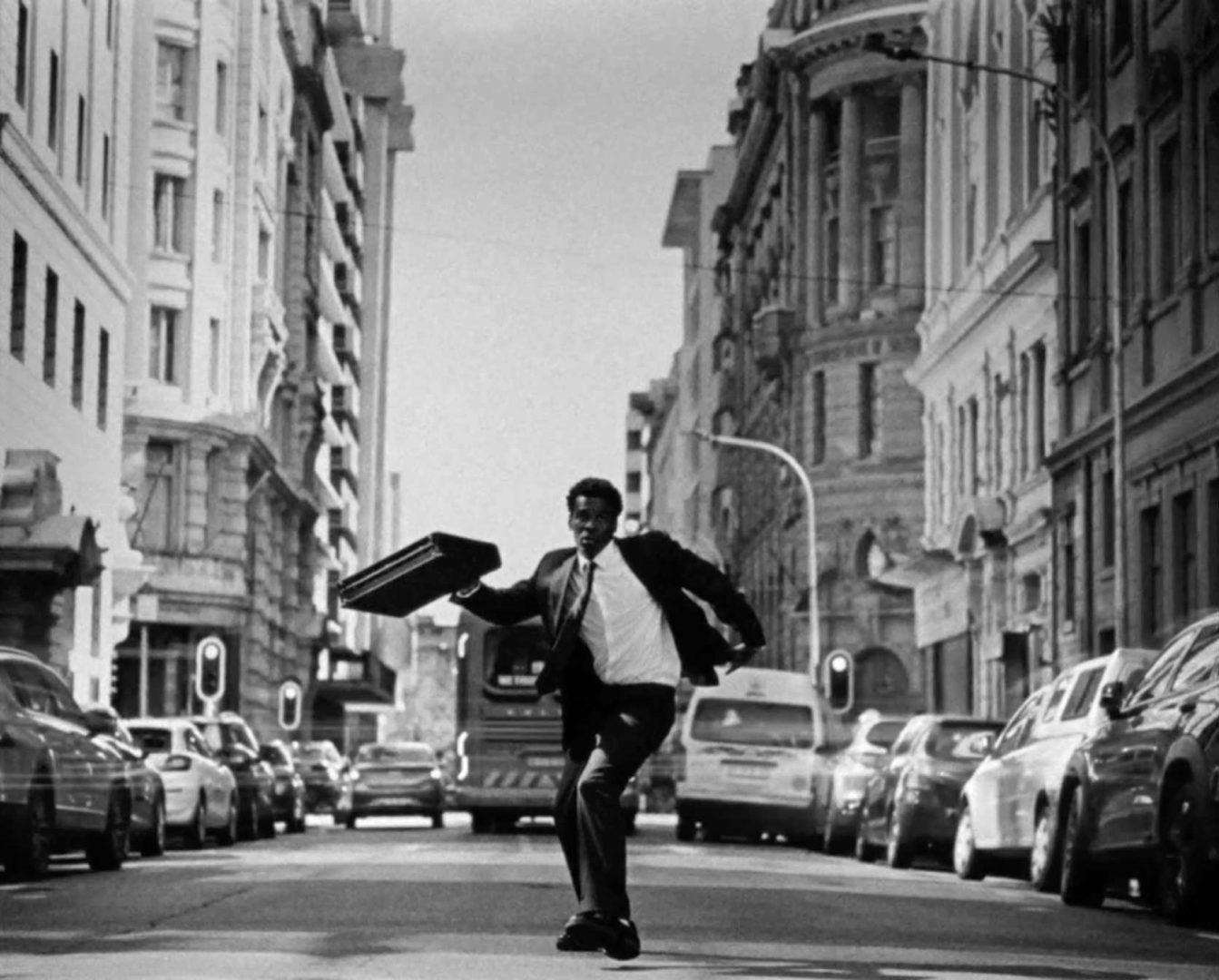 A man in a suit runs down the middle of a busy city street, holding a briefcase—a scene reminiscent of Zee Ntuli’s "African Time." Cars are parked along both sides, and tall buildings line the street. The image, rendered in black and white, captures the urgent pace of urban life. Frame from Stash Magazine article.