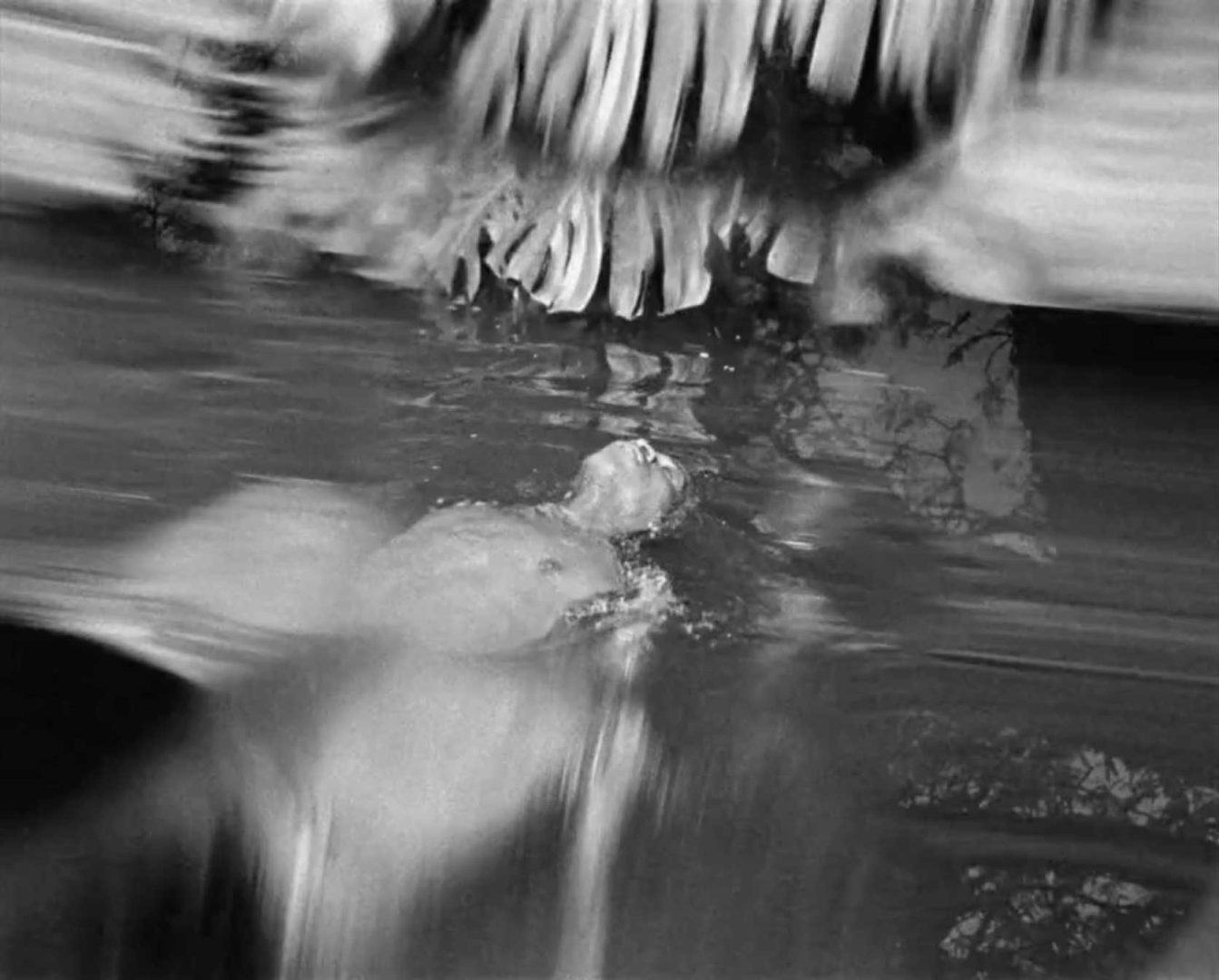 A person floats on their back in calm water, head tilted upward. The scene is blurred, with distorted reflections of white shapes above and abstract light and shadows on the water’s surface—evoking the dreamlike atmosphere seen in the Castle Milk Stout African Time commercial by Zee Ntuli. Frame from Stash Magazine article.