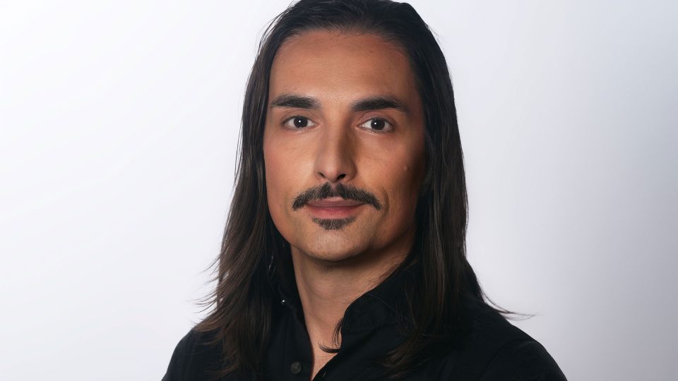A person with long dark hair, a trimmed mustache, and a goatee—resembling Chris-Bahry_CEO_Forever—wears a black collared shirt and poses against a plain light background. Frame from Stash Magazine article.