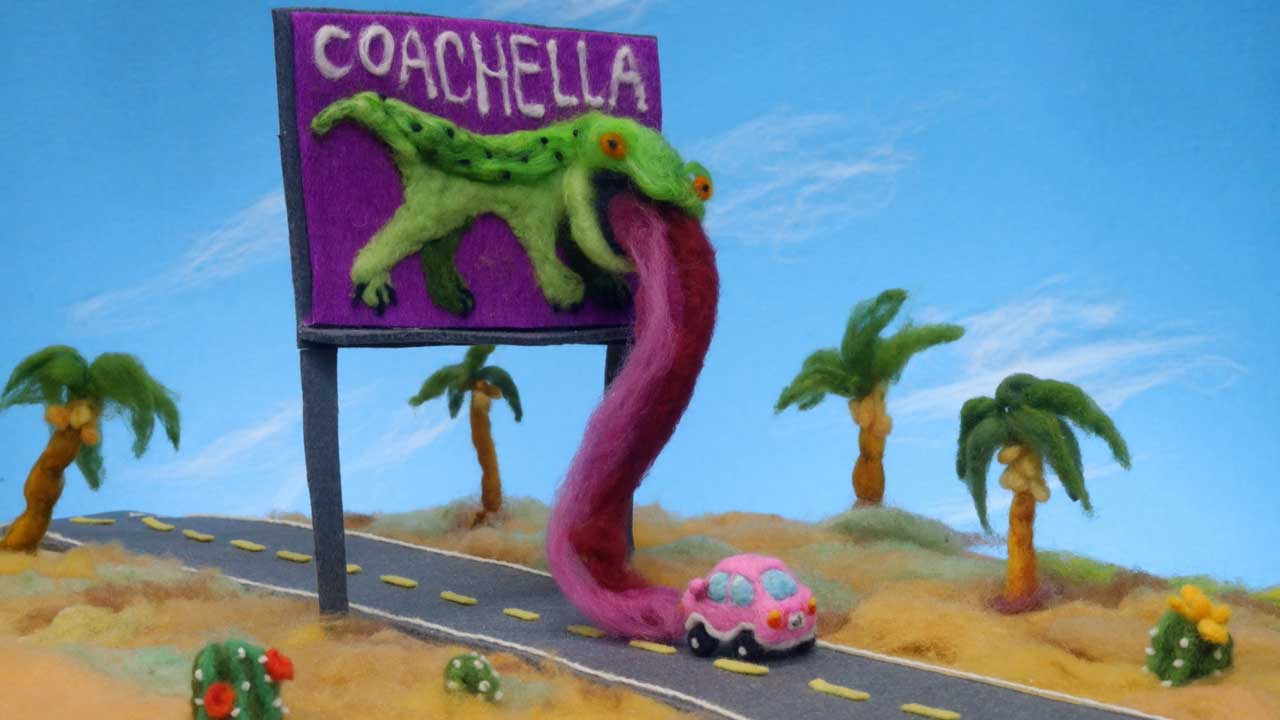 A felt sculpture by Andrea Love depicts a highway scene with a purple Coachella 2026 billboard and a green lizard whose pink tongue reaches toward a small pink car, surrounded by palm trees, desert landscape, and a soaring hornet under the blue sky. Frame from Stash Magazine article.